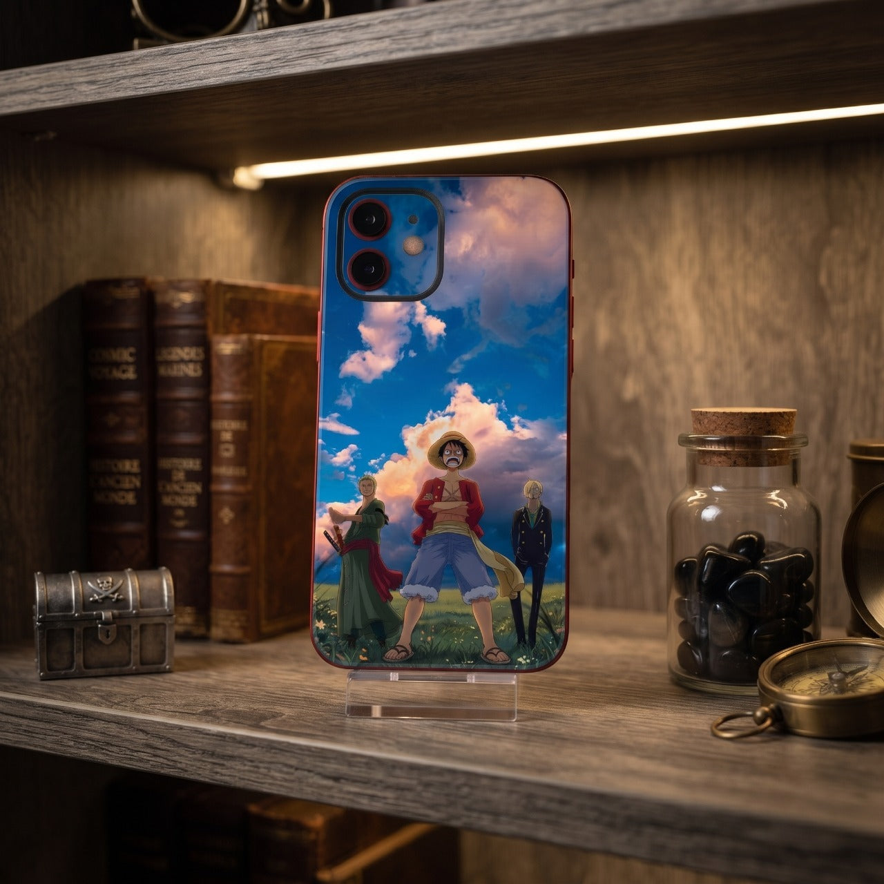 Coque iPhone One Piece Luffy Zoro et Sanji sur un support, étagère en bois avec livres anciens, coffre au trésor et boussole