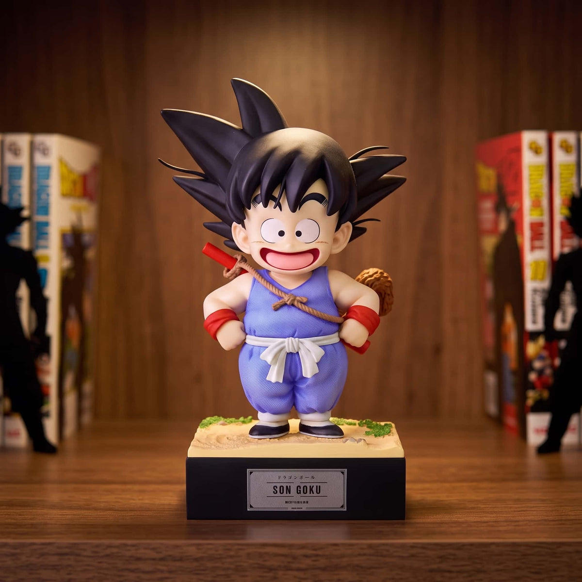 Figurine de Goku enfant de 17 cm, gi bleu et bâton magique dans le dos, queue de singe visible, sur un socle noir à plaque posé sur une étagère en bois