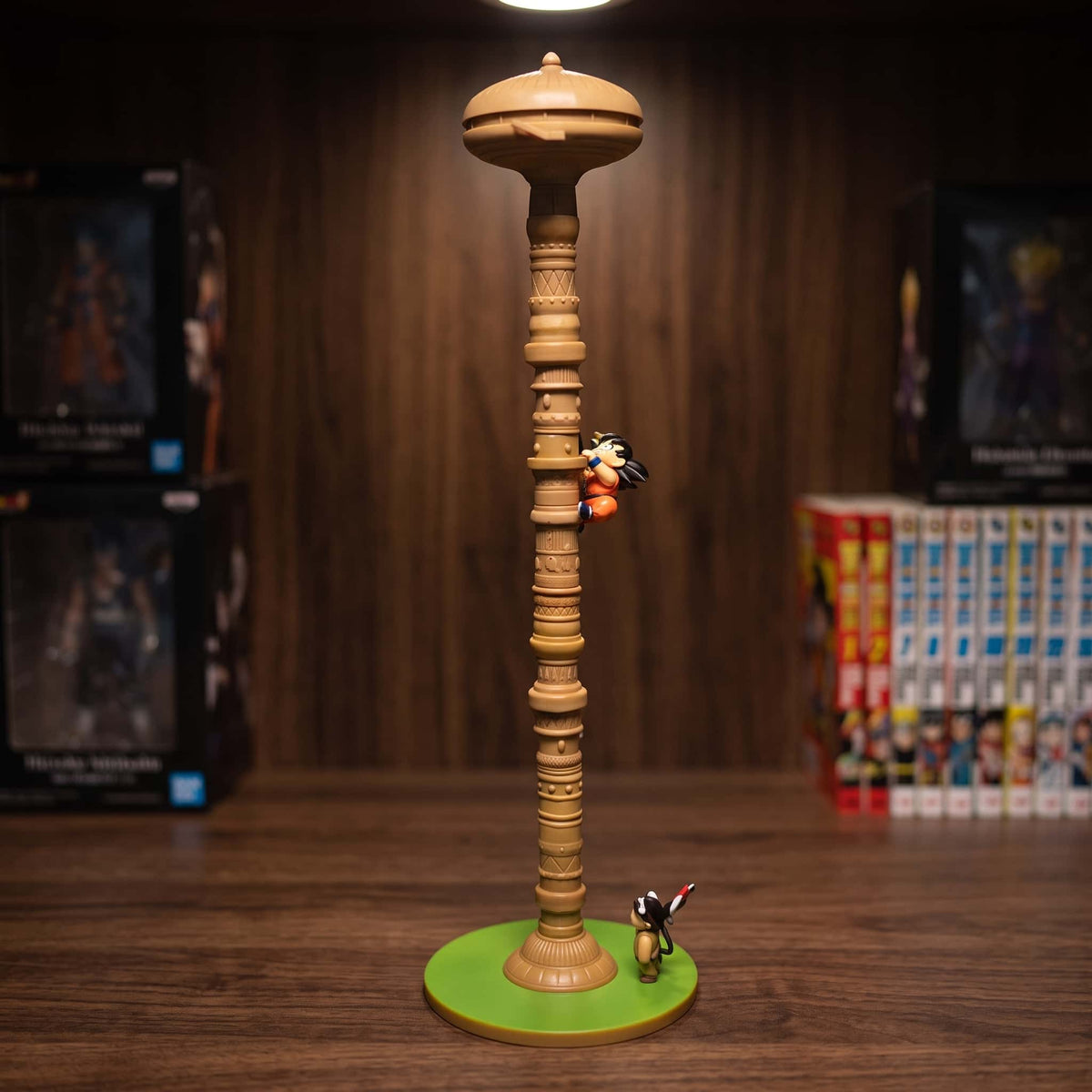Figurine de la Tour de Karin de Dragon Ball : colonne beige annelée de 35 cm coiffée d'un disque en champignon, Son Goku enfant agrippé à mi-hauteur et Upa au pied, socle d'herbe verte, posée sur une étagère en bois