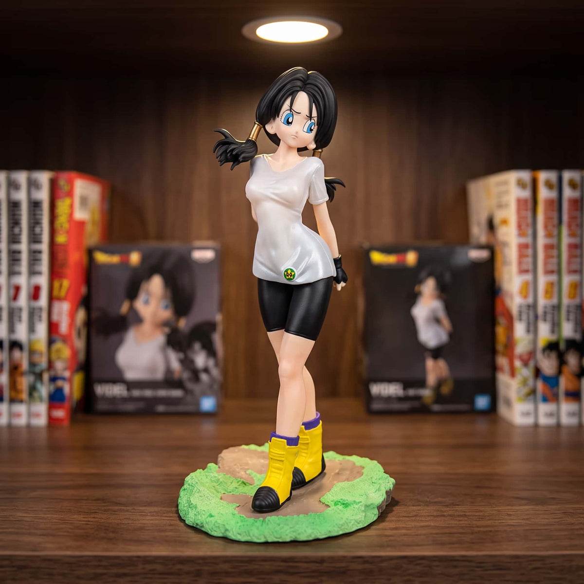 Figurine de Videl de Dragon Ball Z : t-shirt blanc, cycliste noir, mitaines et bottes jaunes à revers violet, deux couettes basses, 25 cm sur un socle d'herbe verte, posée sur une étagère en bois