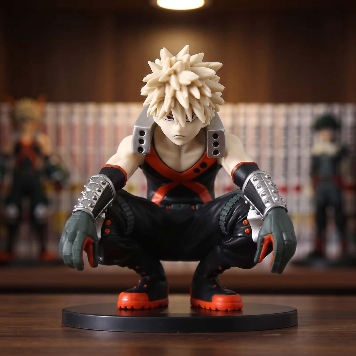 Figurine de Katsuki Bakugo accroupi, débardeur noir à X orange et gantelets argentés, 11 cm sur socle noir, posée sur une étagère en bois