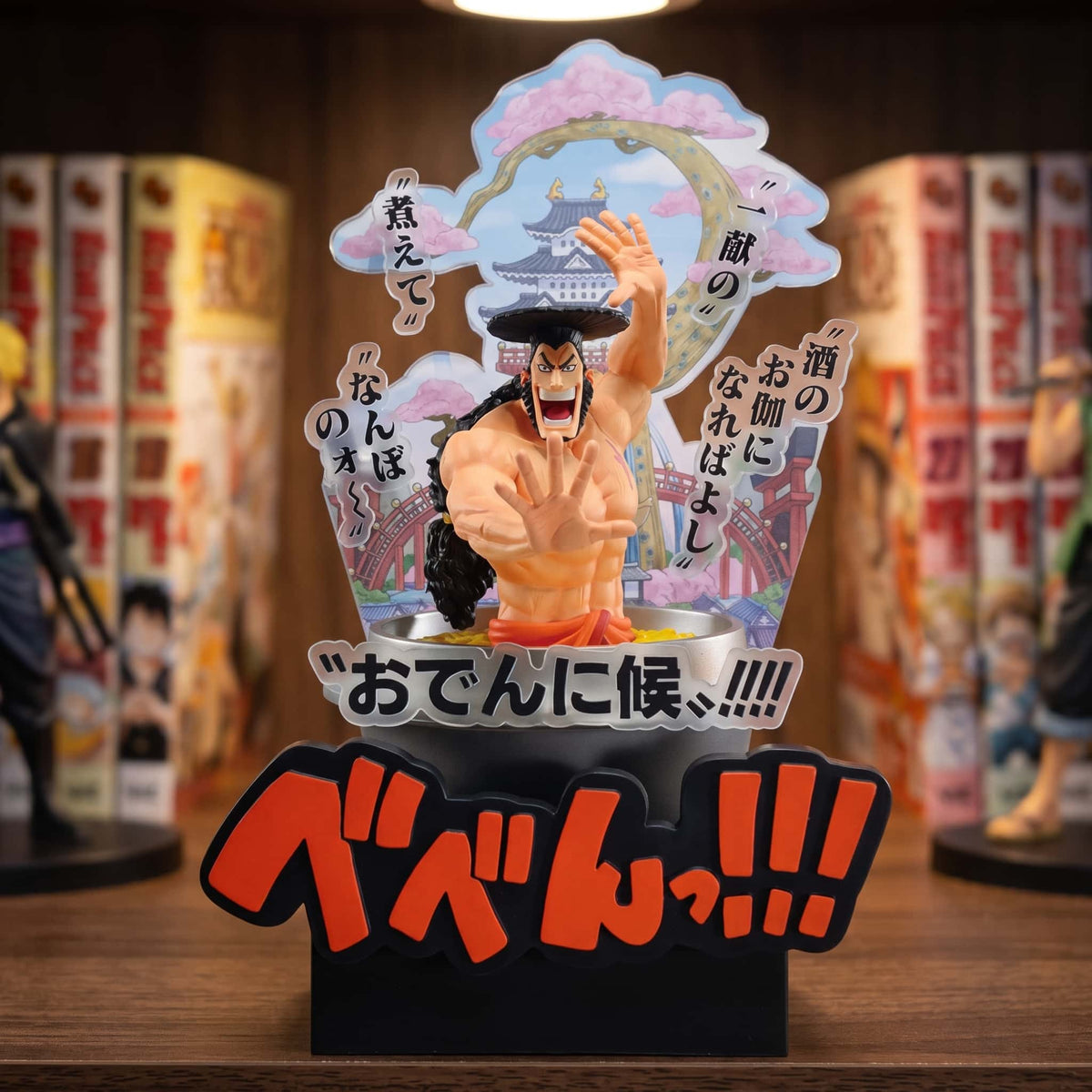 Figurine One Piece Kozuki Oden 22 cm dans son chaudron devant un panneau illustré de Wano, exposée sur une étagère entre des mangas (décor non inclus)