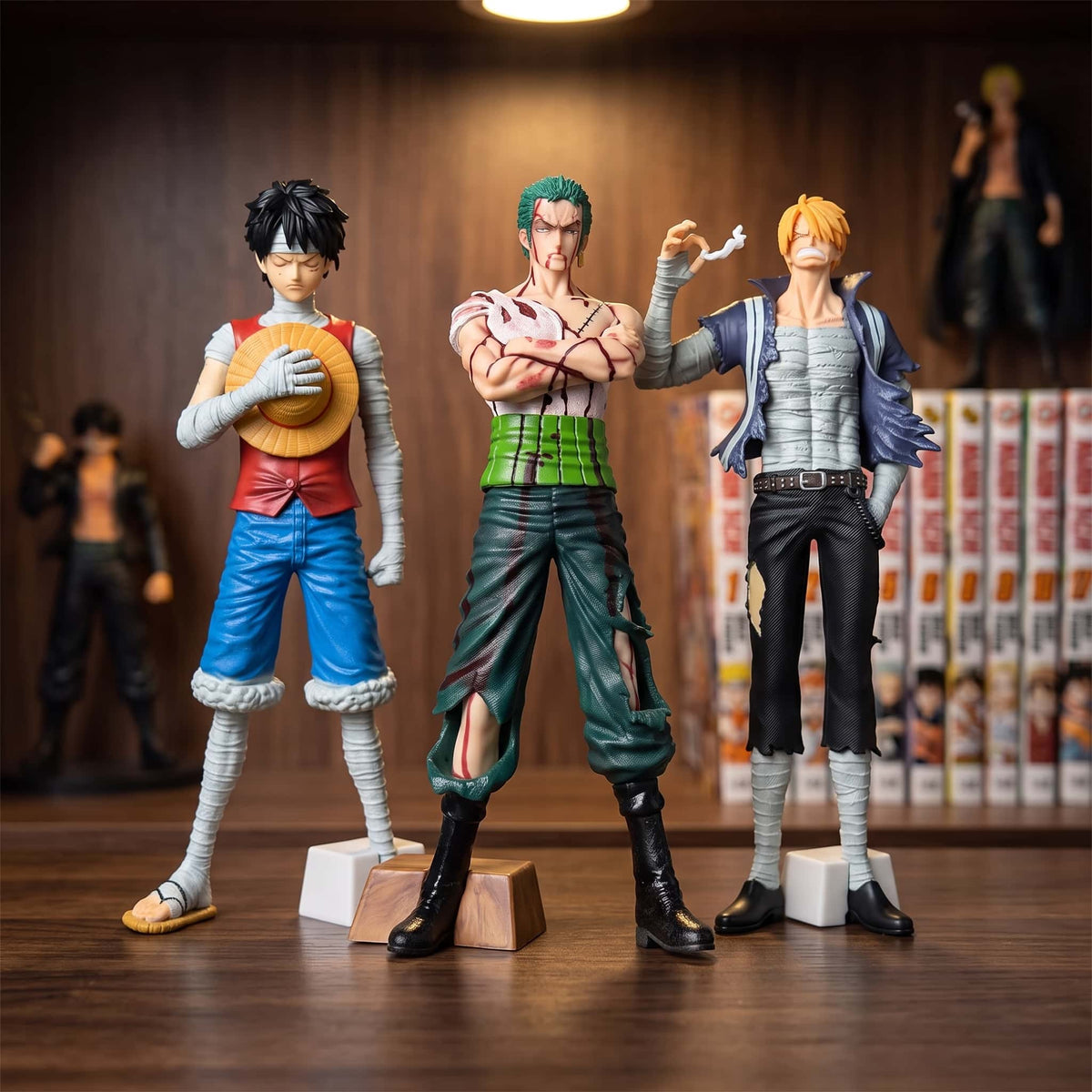 Figurines One Piece Luffy et Sanji couverts de bandages et Zoro ensanglanté, debout sur une étagère devant des mangas (vendues à l'unité, décor non inclus)