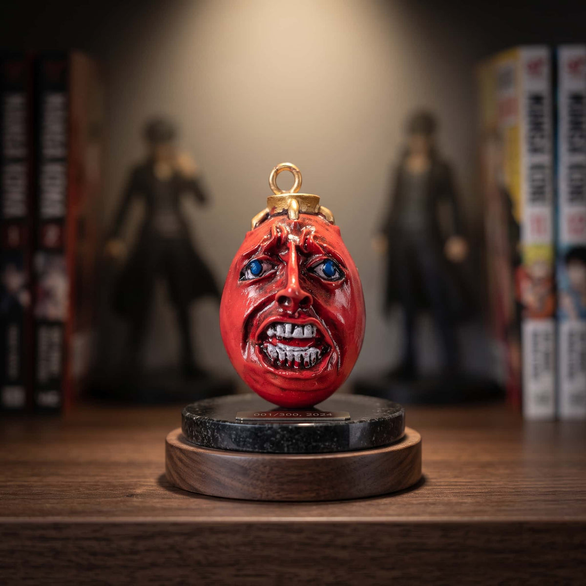 Pendentif Behelit de Berserk : pierre rouge sculptée d'un visage humain aux yeux bleus, bélière dorée, posé sur un socle de présentation devant des tomes de manga