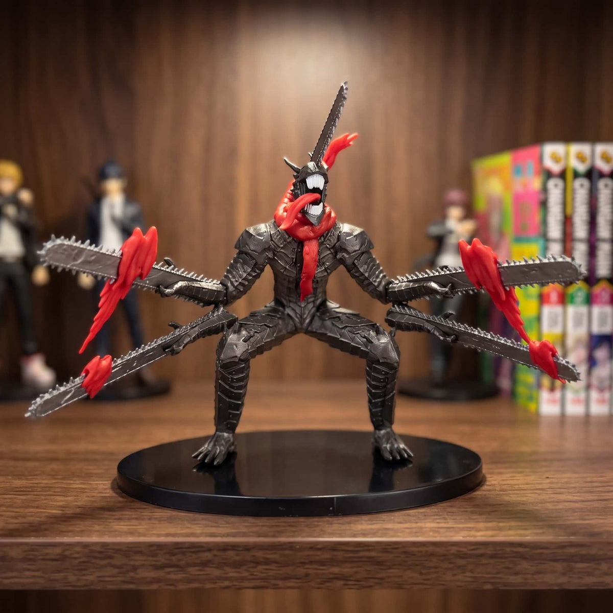 Figurine du Démon Tronçonneuse de Chainsaw Man : corps noir écaillé, cinq lames sorties du crâne, des bras et des cuisses, langue rouge pendante, 18 cm sur socle noir, posée sur une étagère en bois