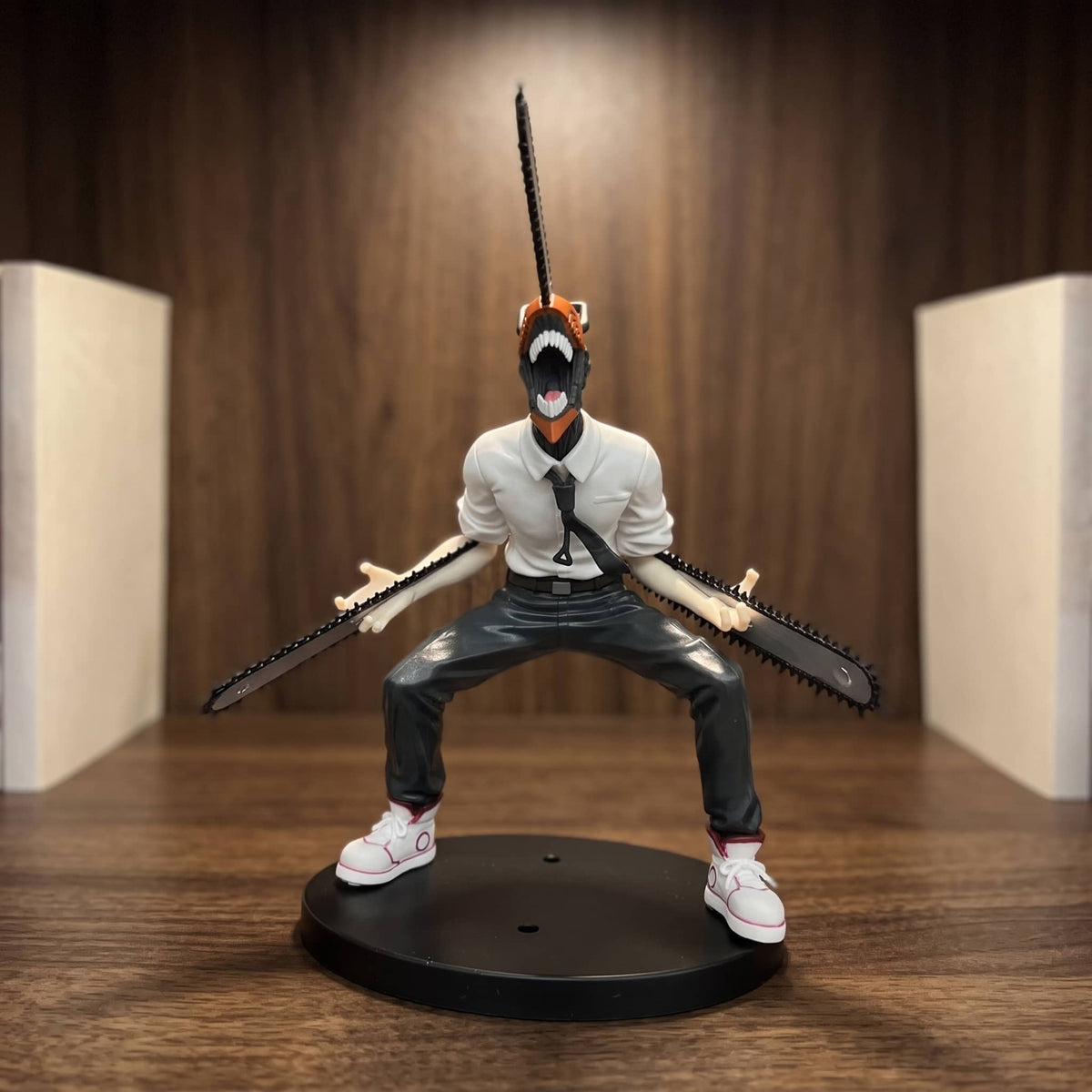 Figurine de Denji transformé en Chainsaw Man : tête-tronçonneuse orange et noire gueule ouverte, lames sorties des deux avant-bras, chemise blanche et cravate, 19 cm sur socle noir, posée sur une étagère en bois