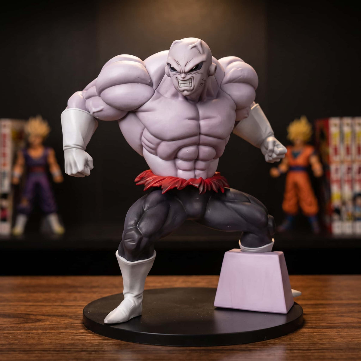 Figurine de Jiren pleine puissance : buste cambré en avant, musculature exagérée, peau violette et ceinture rouge effilochée, un pied posé sur un bloc mauve, 25 cm sur socle noir, posée sur une étagère en bois