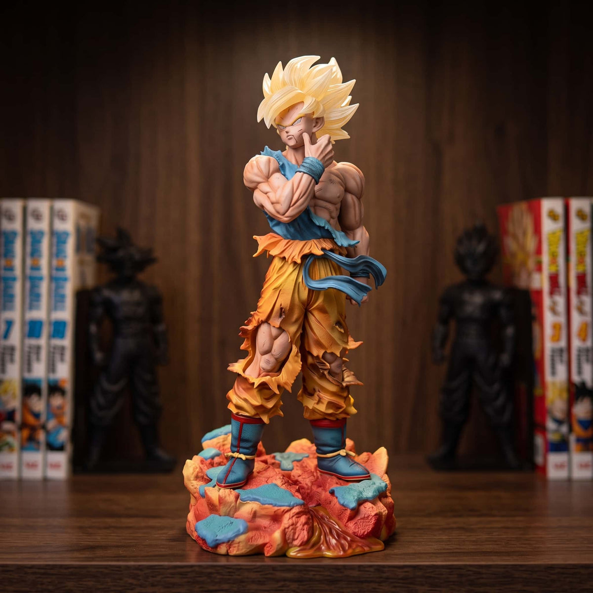 Figurine de Son Goku Super Saiyan bras croisés : mèche blonde sur le front, haut bleu déchiré et bas de gi orange en lambeaux, 37 cm sur un socle de rochers orange et bleus, posée sur une étagère en bois