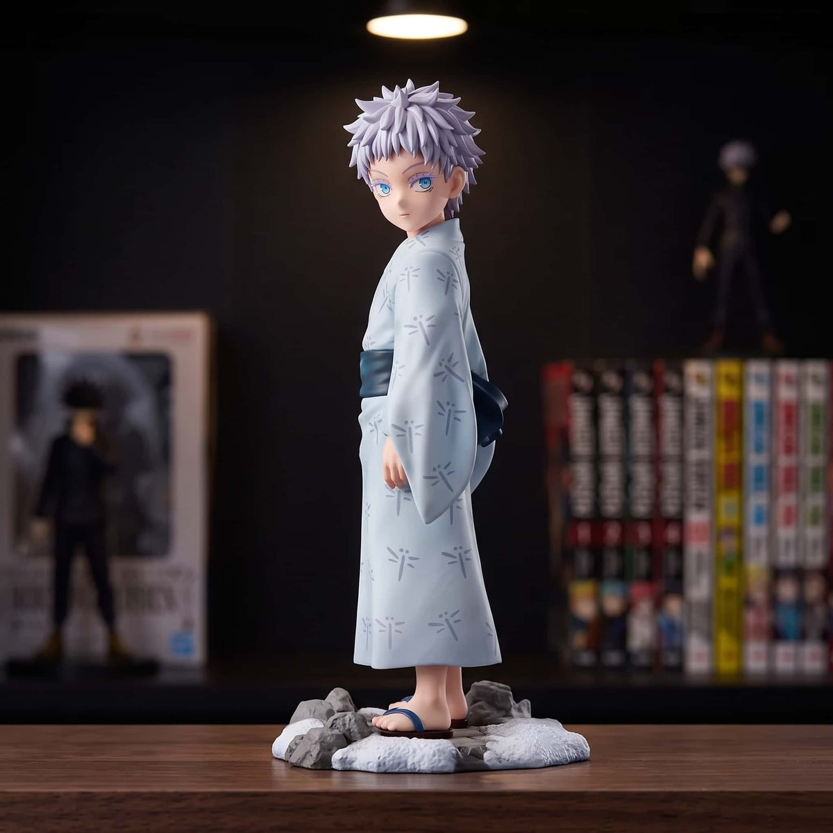 Figurine de Gojo Satoru enfant en yukata bleu pâle à motifs de libellules, obi bleu nuit et geta, sur un socle de rochers enneigés, 14 cm, posée sur une étagère en bois sombre