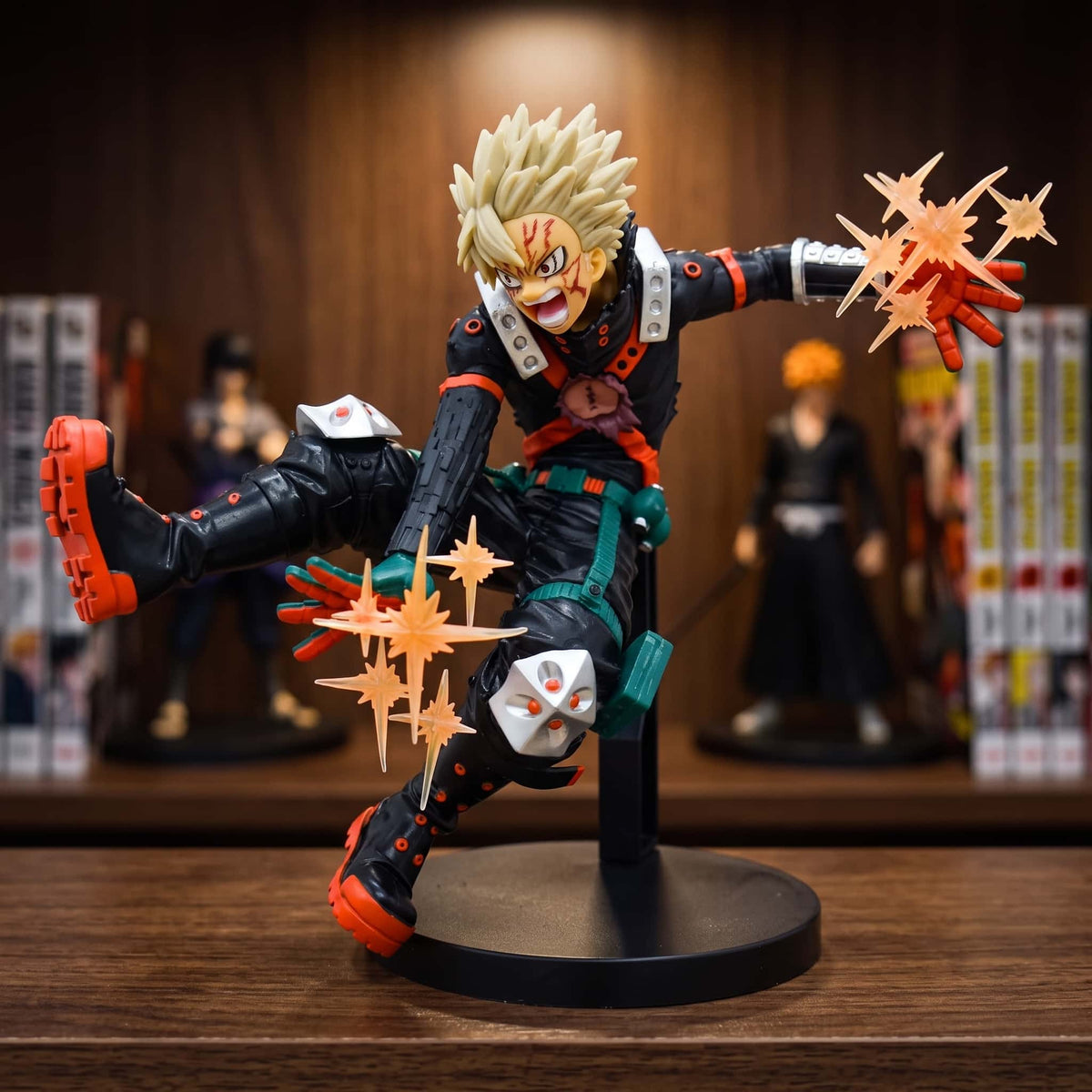 Figurine de Katsuki Bakugo en pleine charge, deux explosions orange translucides aux mains, cicatrice au visage et tenue déchirée, 18 cm sur socle noir, posée sur une étagère en bois