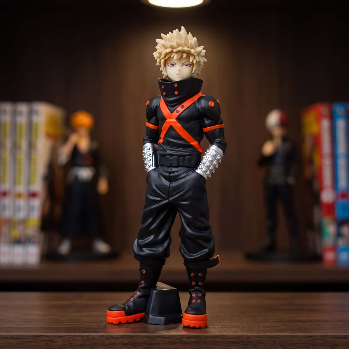 Figurine de Katsuki Bakugo debout, mains dans les poches, combinaison noire à harnais orange et gantelets argentés, 21 cm, posée sur une étagère en bois