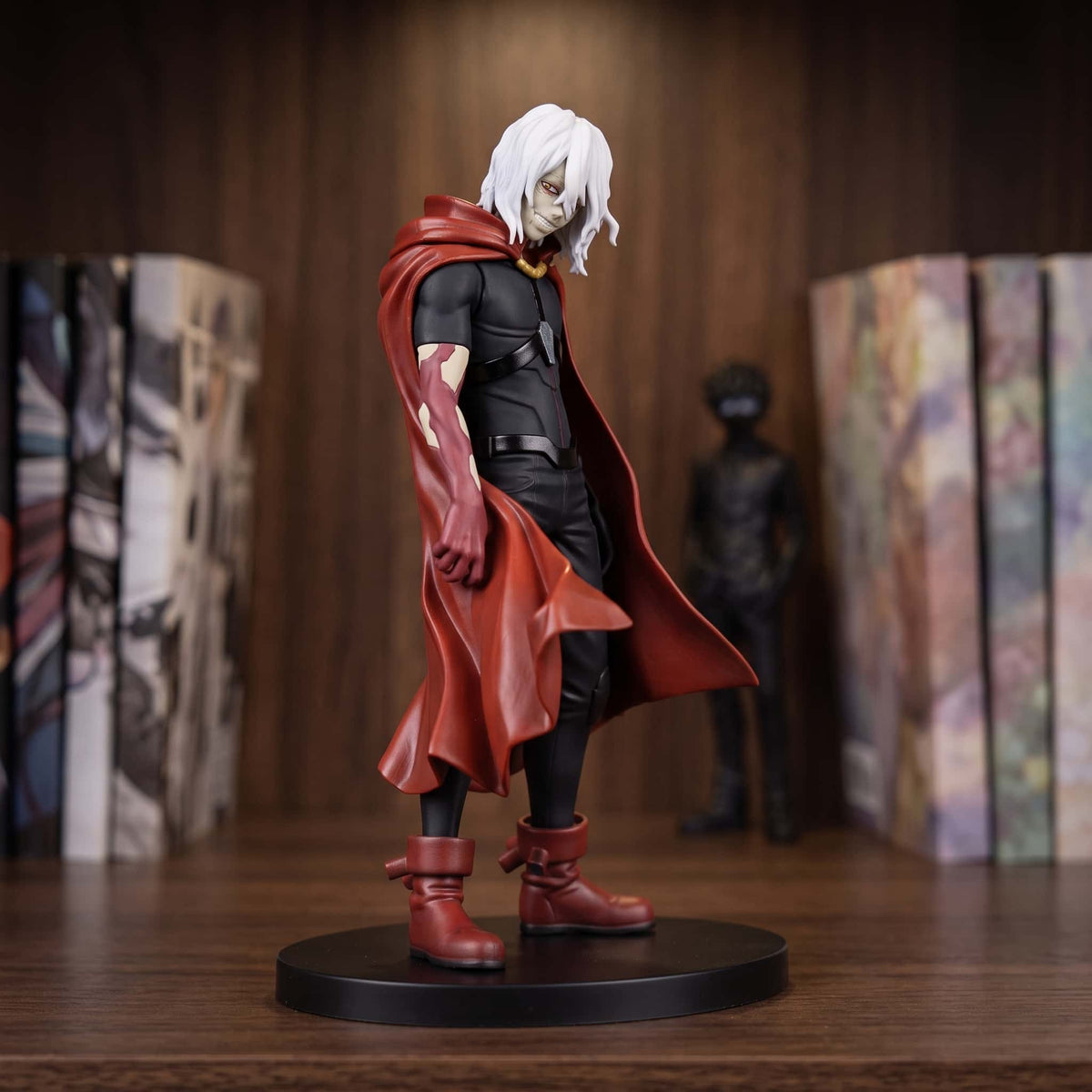 Figurine de Shigaraki Tomura aux cheveux blancs, long manteau rouge ouvert et bottes rouges, sur un socle noir posé sur une étagère en bois