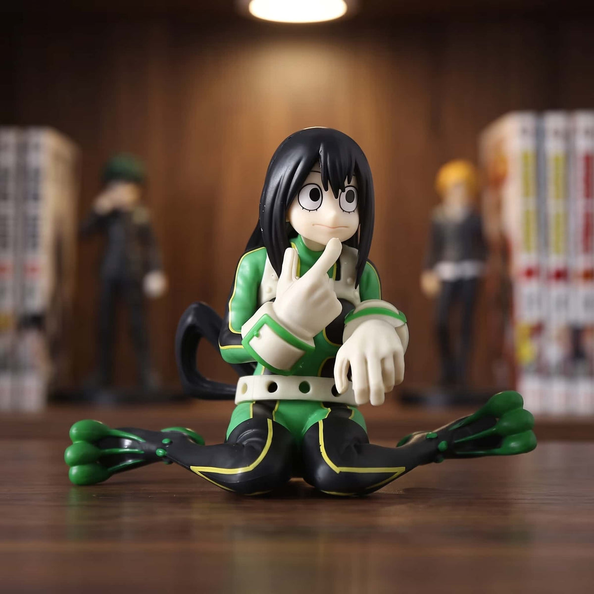 Figurine de Tsuyu Asui assise au sol, index replié contre le menton, combinaison verte et noire et gants palmés, 9 cm, posée sur une étagère en bois