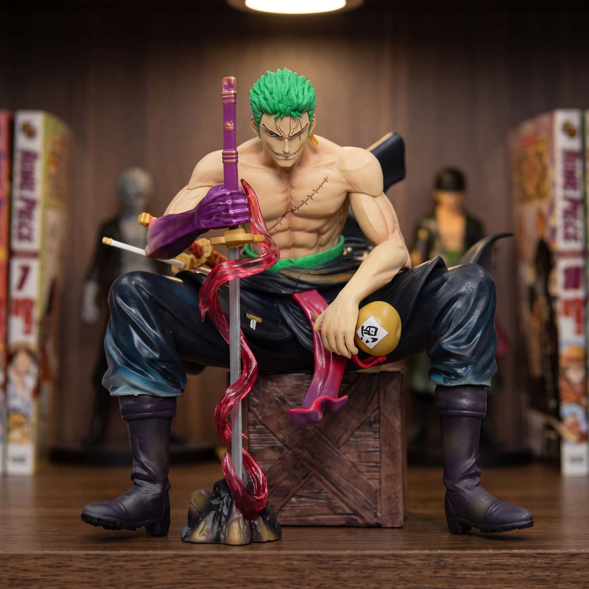 Figurine One Piece Roronoa Zoro 15 cm assis sur une caisse, sabre Enma planté devant lui, exposée sur une étagère entre des mangas (décor non inclus)