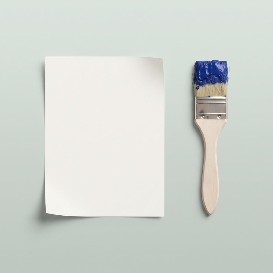 Affiche vierge en papier ivoire posée à plat à côté d'un pinceau chargé de peinture bleue, fond vert doux