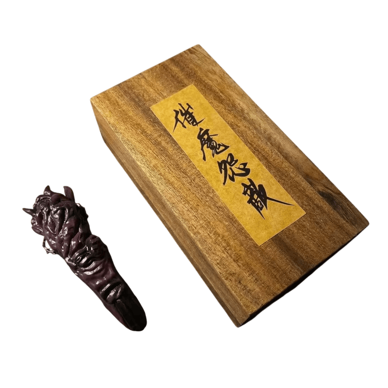 Réplique du doigt de Sukuna posée à plat à côté de sa planche de bois, vue de dessus : doigt brun-aubergine à base noueuse et étiquette de papier calligraphiée